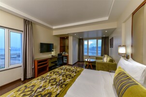 A hotel room with a large bed, a desk with a TV, a small table, a chair, and a view of the city through large windows.