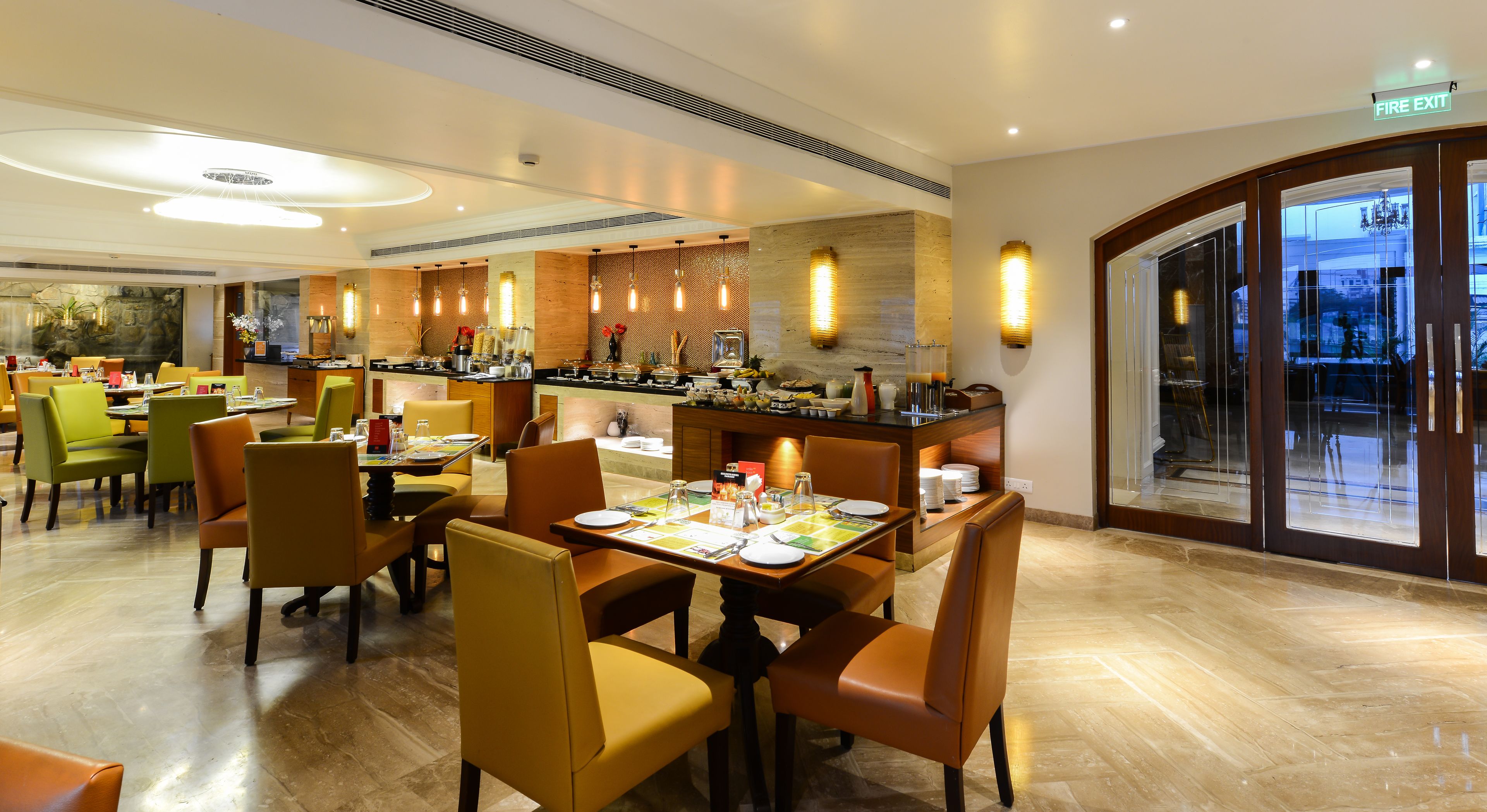 A hotel dining area with multiple tables and chairs, a kitchen counter, and a fire exit sign.