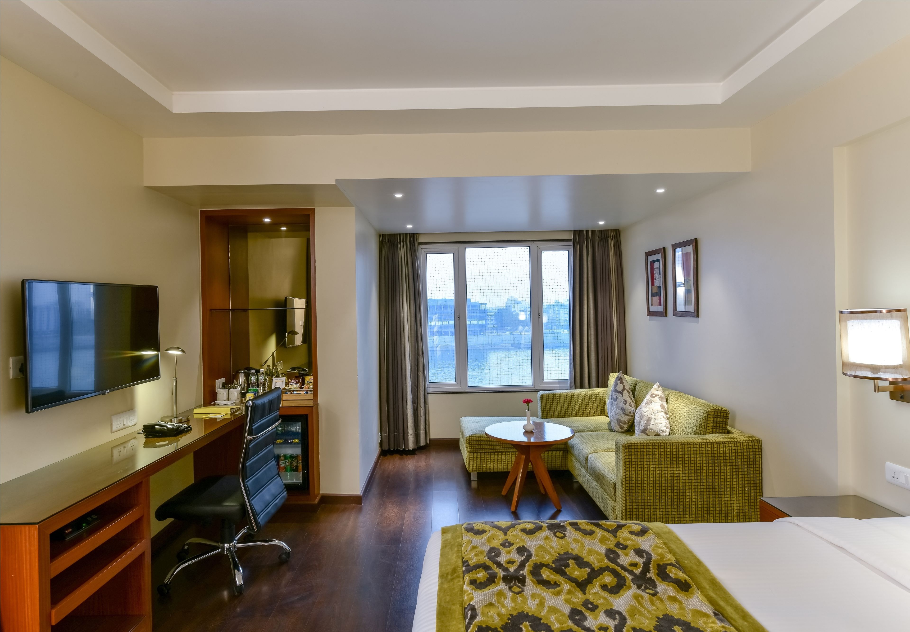 Une chambre d’hôtel avec un lit, un bureau avec un ordinateur, une télévision, un canapé et une fenêtre donnant sur la ville.
