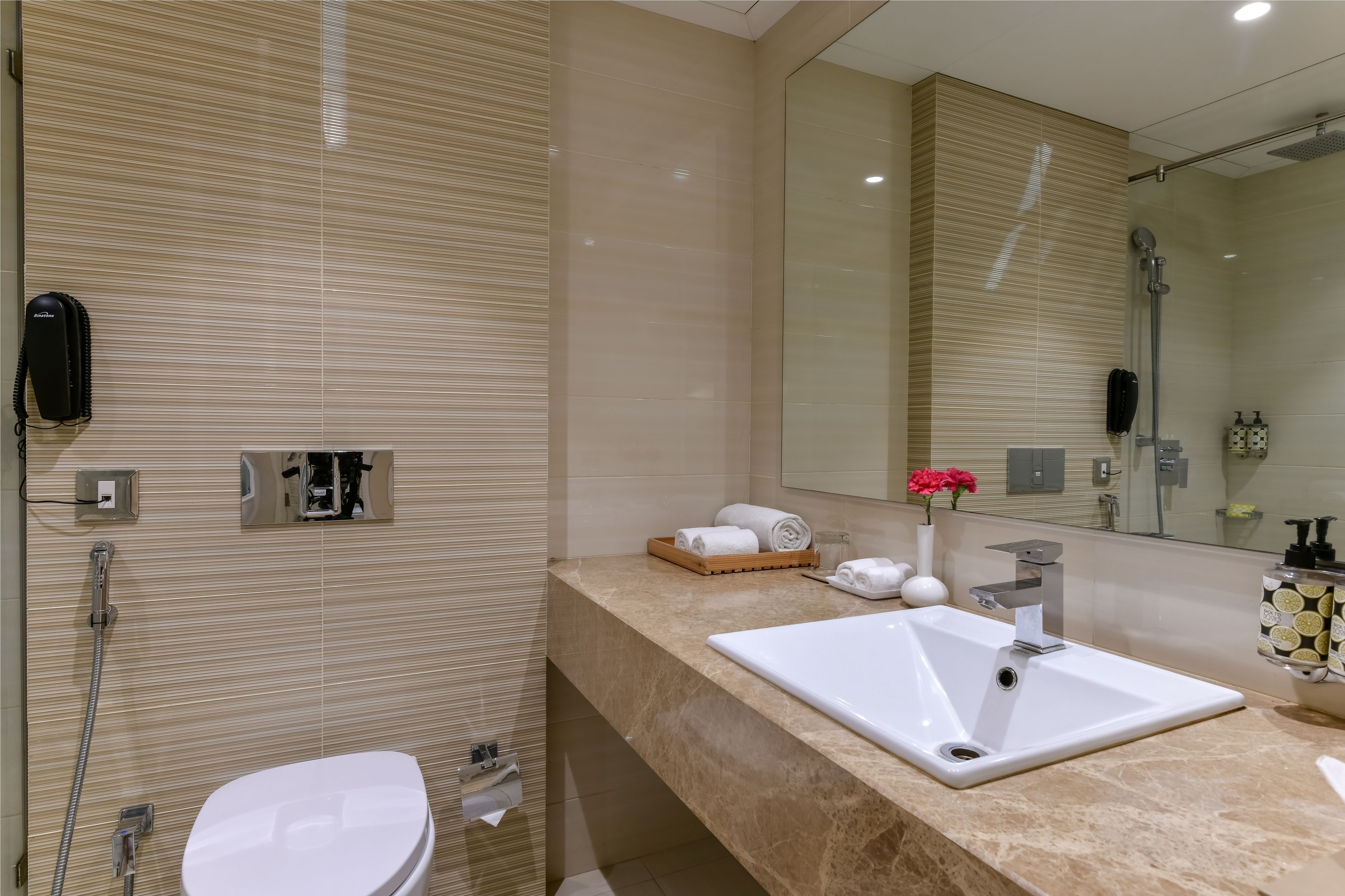A modern bathroom with a beige tiled wall, a white sink, a toilet, a shower, and a large mirror.