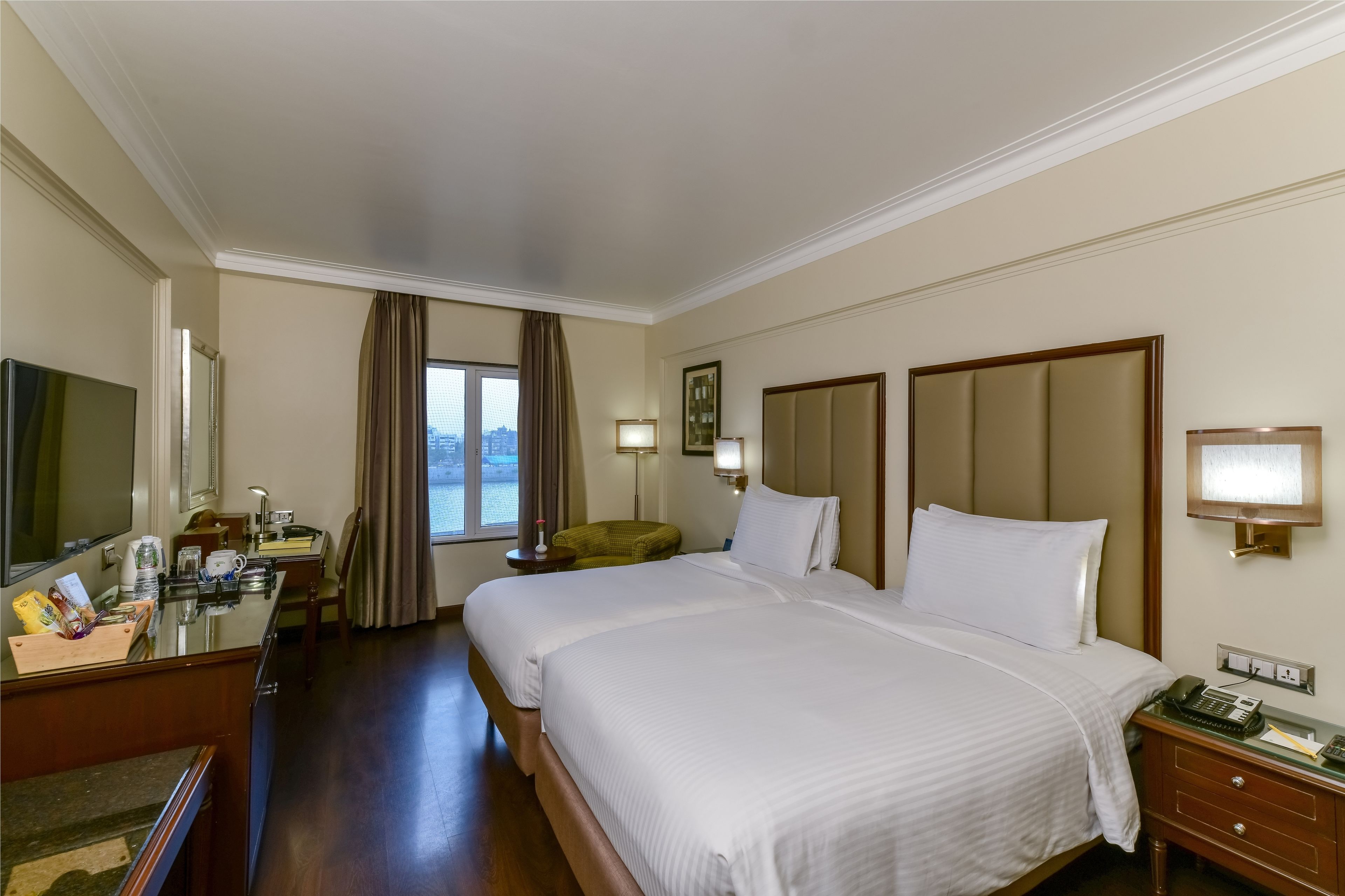 Quarto de hotel com duas camas, uma escrivaninha, uma cadeira, televisão e janela com cortinas.