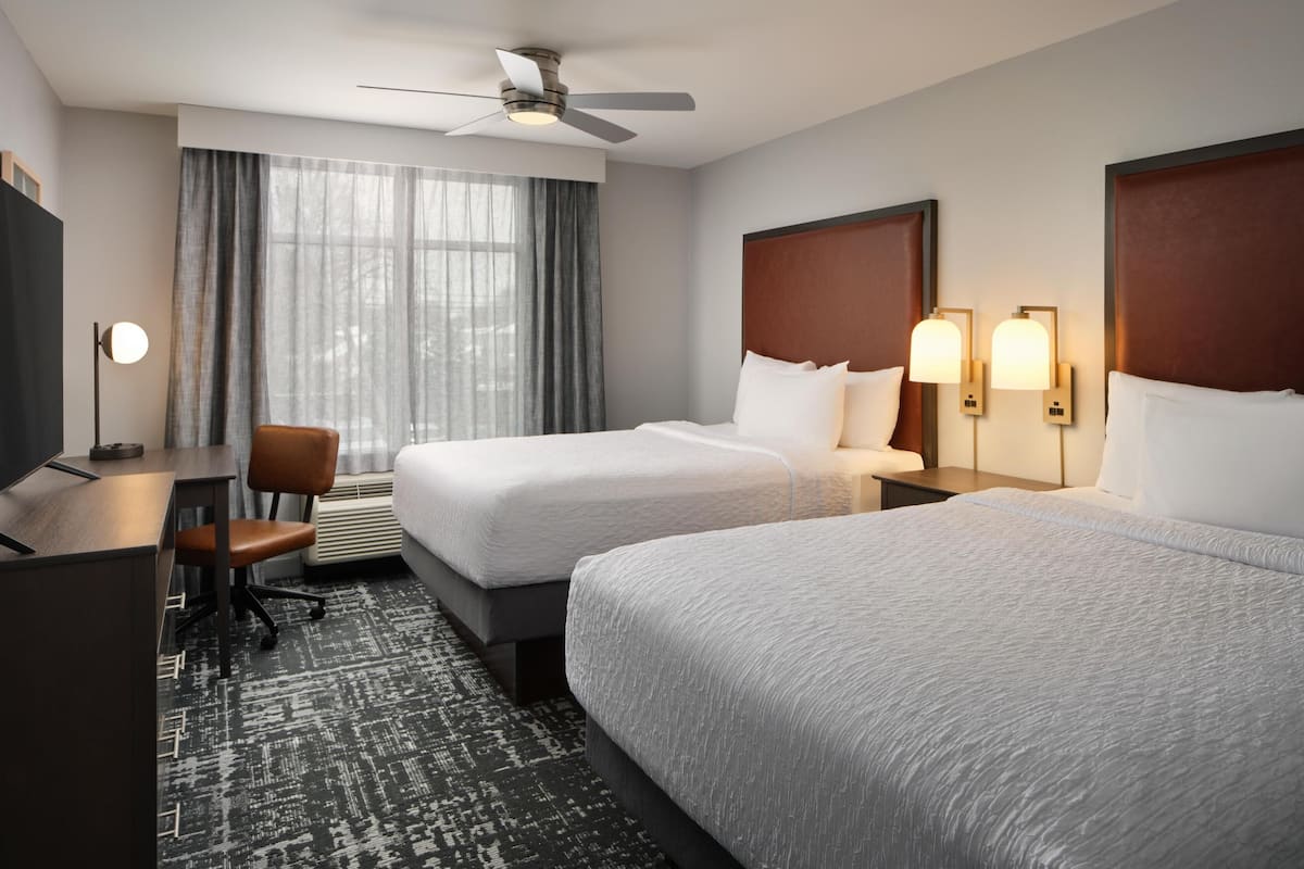 A hotel room with two beds, a desk, a chair, a ceiling fan, and a window with curtains.