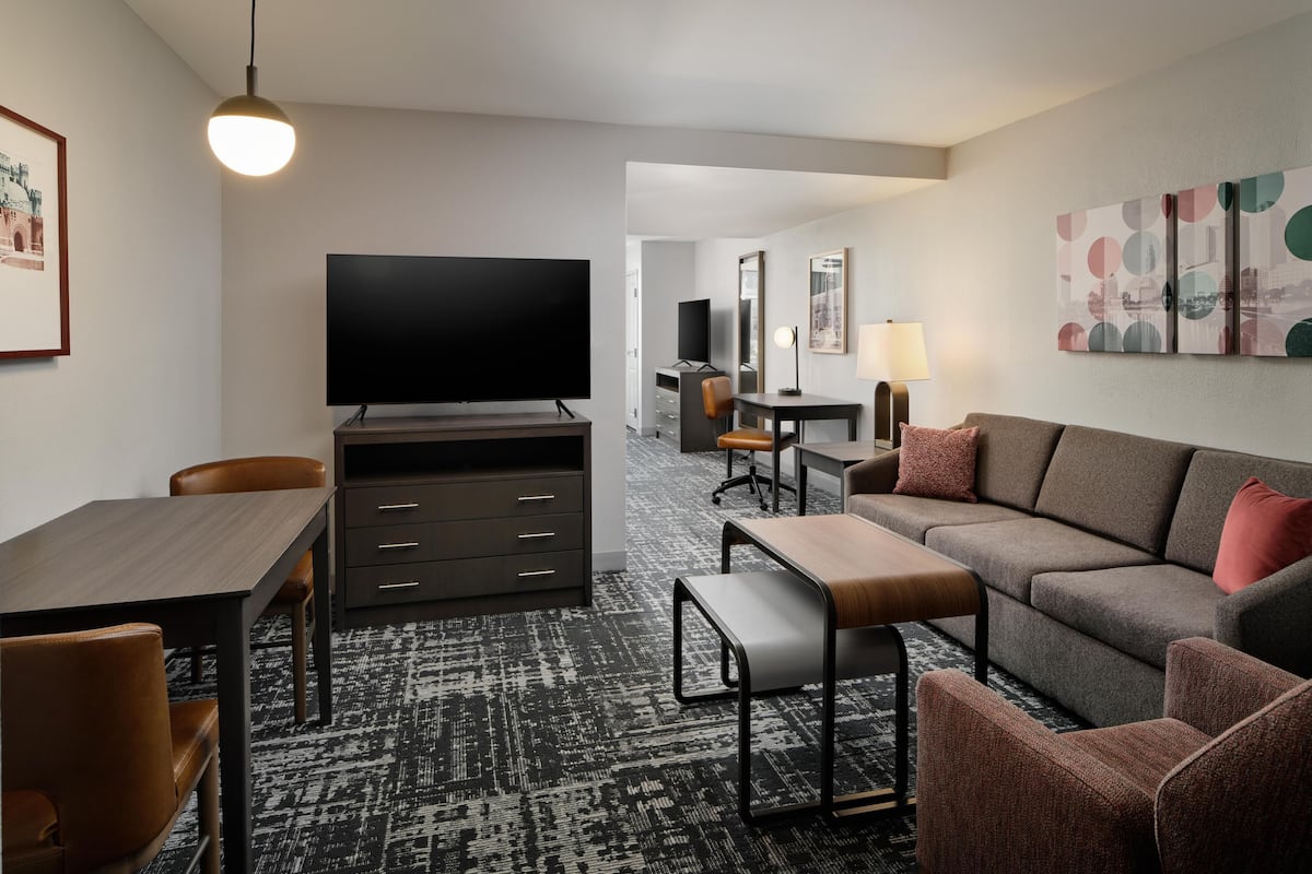 A modern hotel room with a flat-screen TV, a sofa, a desk with a chair, and a lamp.