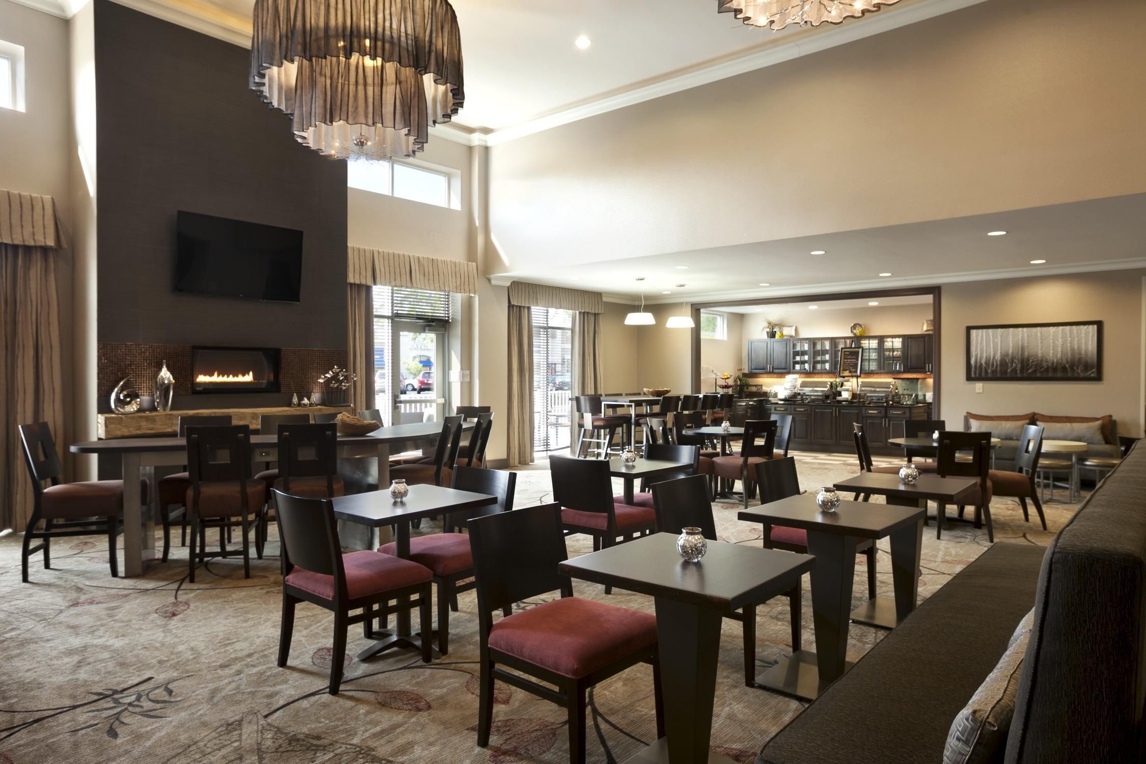 A hotel lobby with multiple seating areas, a fireplace, a television, and a kitchen in the background.