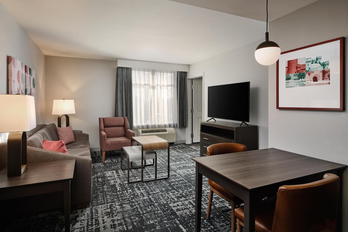 A hotel room with a sofa, armchair, small table, TV, and a framed picture on the wall.