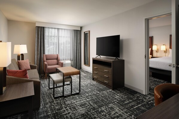 Quarto de hotel com sofá, poltrona, televisão e cama com mesa de cabeceira.