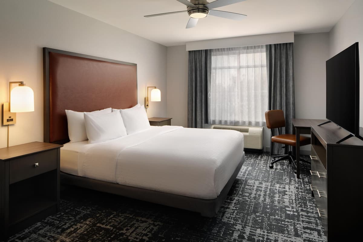A hotel room with a bed, a desk, a chair, a TV, a ceiling fan, and a window with curtains.