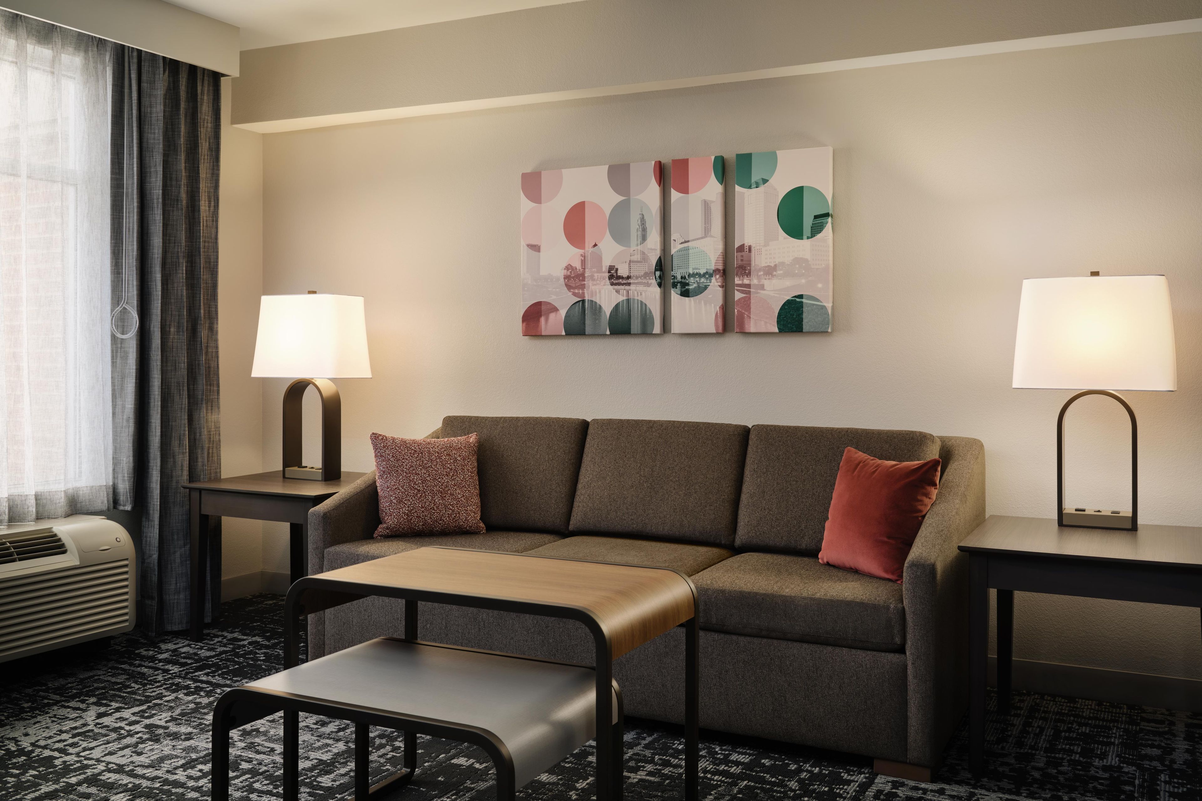 A hotel room with a sofa, two side tables with lamps, a small table, a chair, and a large abstract painting on the wall.