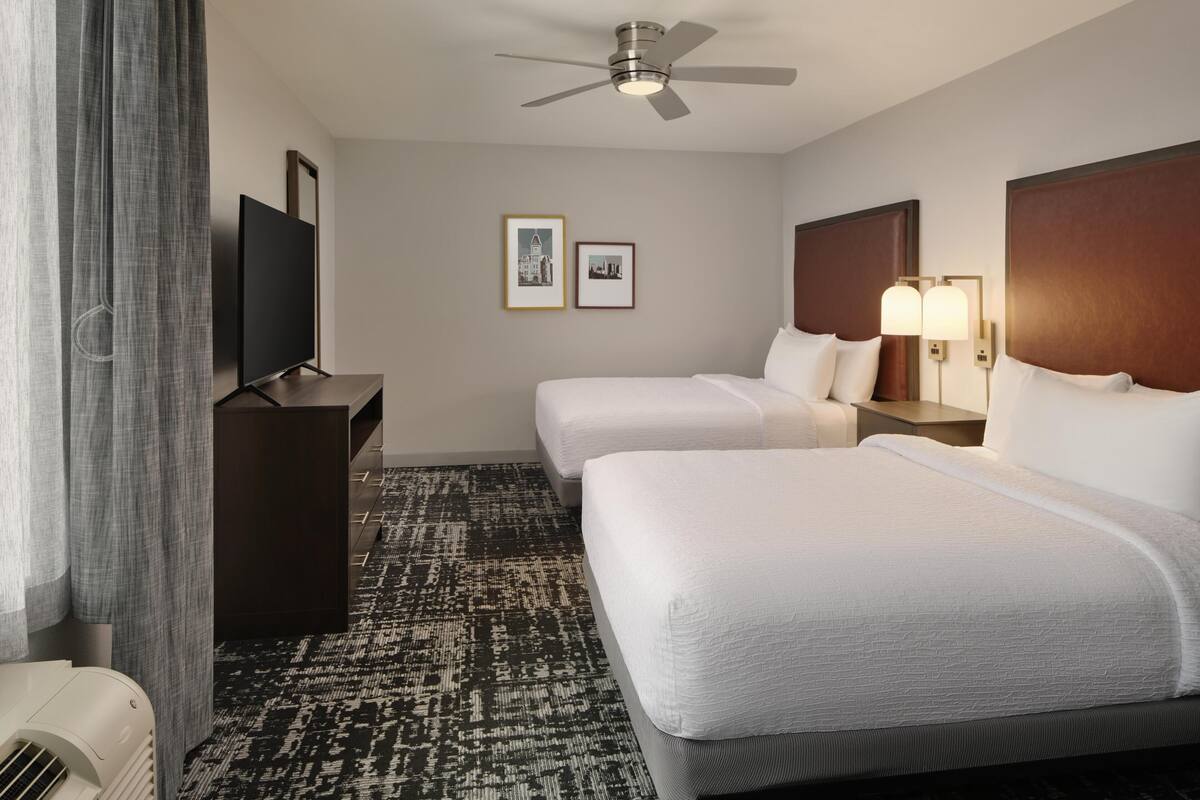 A hotel room with two beds, a flat-screen TV, a ceiling fan, framed pictures on the wall, and a patterned carpet.