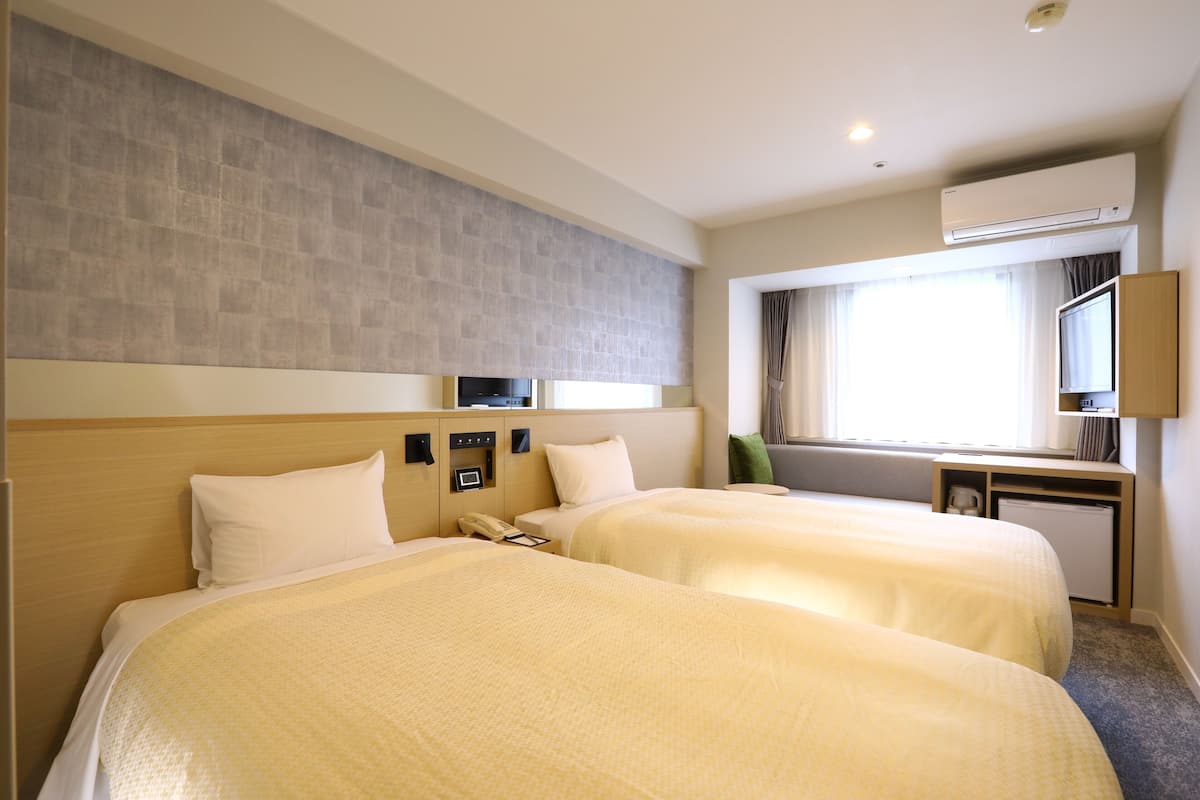 Une chambre d’hôtel avec deux lits, un bureau, une chaise, un climatiseur et une fenêtre avec des rideaux.