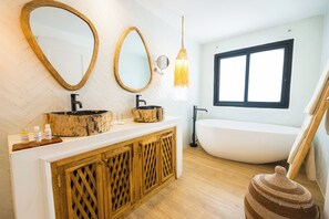 Un baño moderno con un tocador de madera, dos espejos, una bañera independiente y un ventanal amplio.