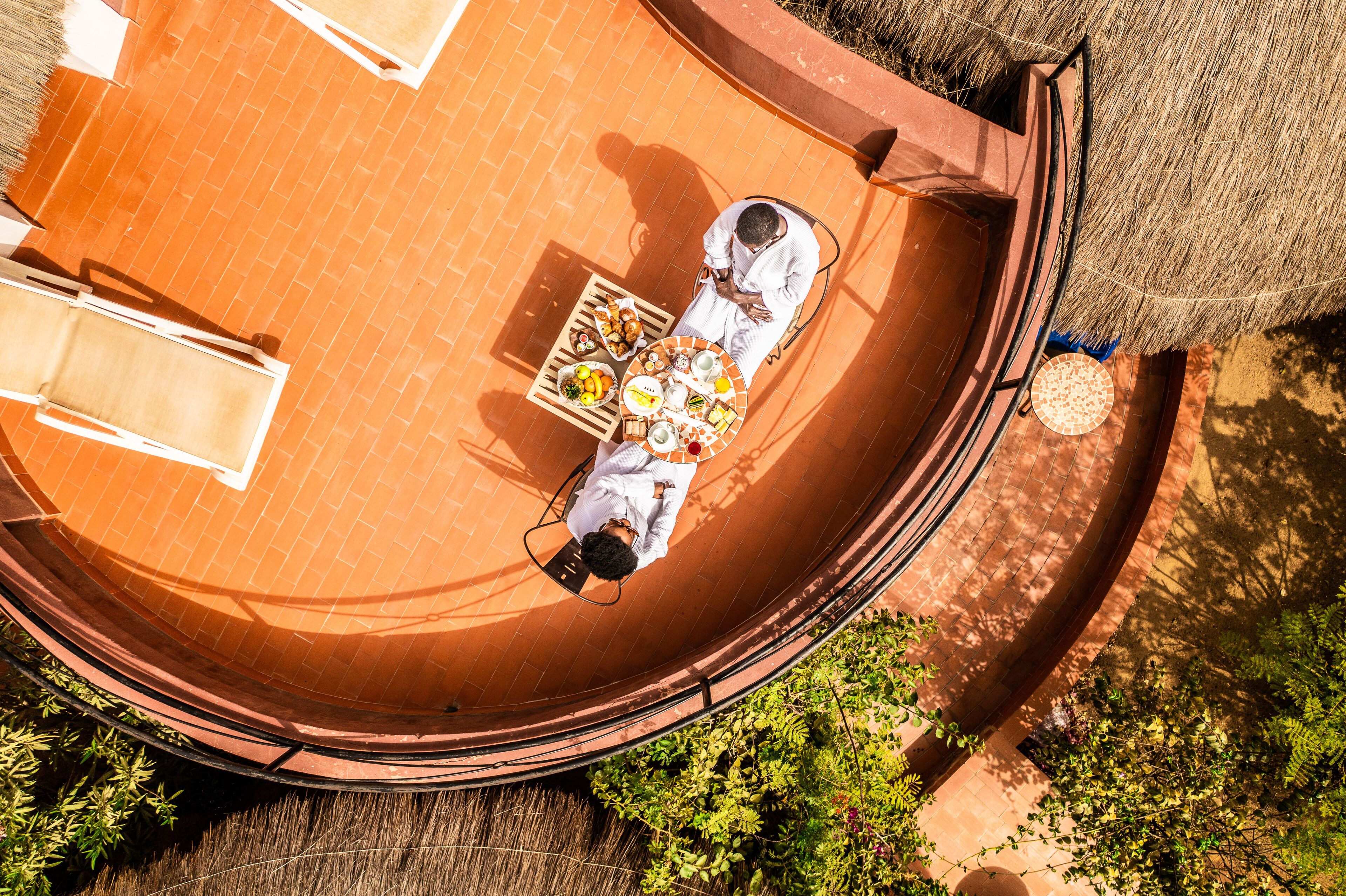 An aerial view of a circular outdoor dining area with a round table set for two, surrounded by a brick floor and greenery.