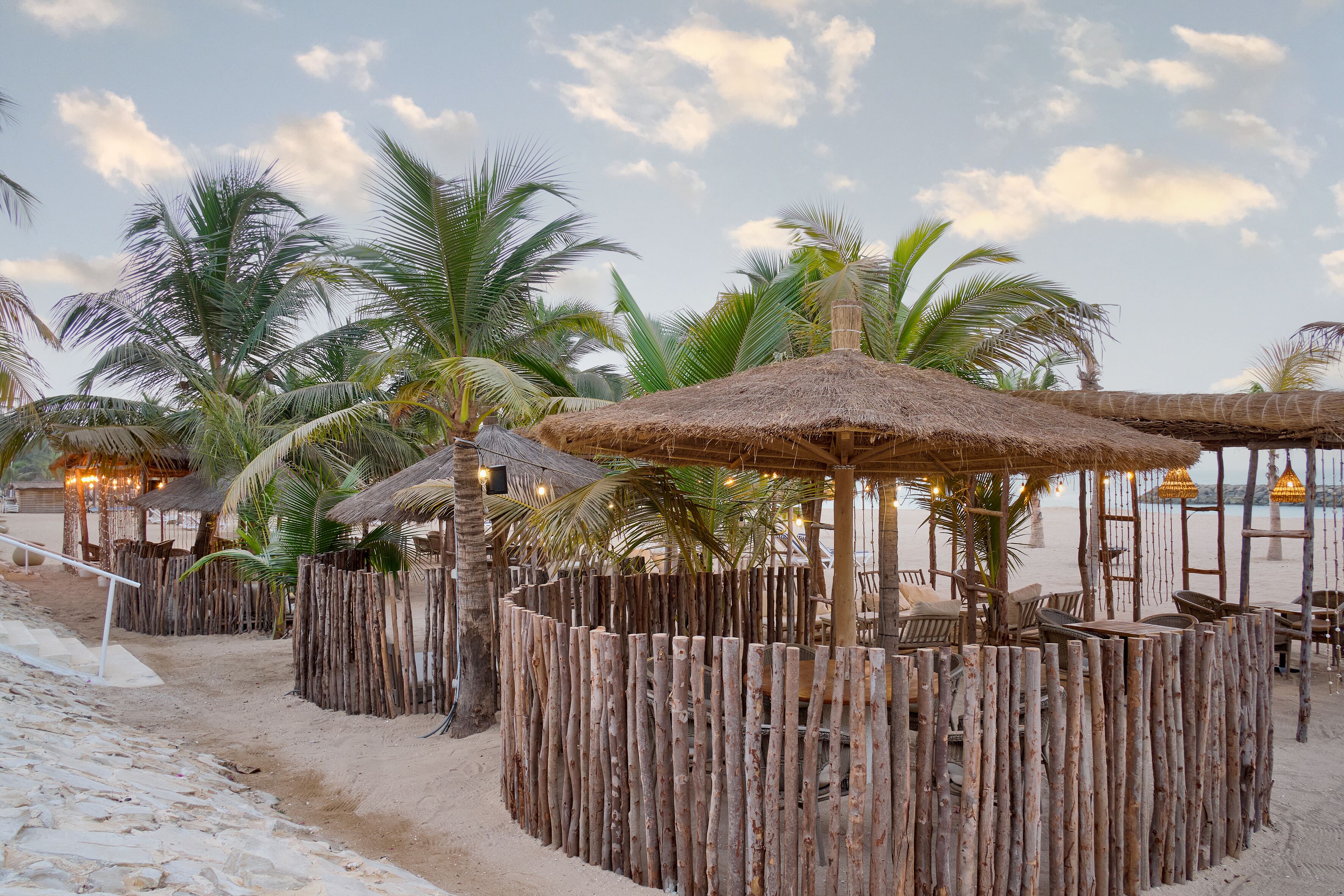 A beachside area with thatched-roof structures, palm trees, and sandy ground.
