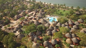 Vista aérea de un resort con cabañas de techo de paja, una piscina central y una playa en el fondo.