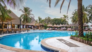 Una piscina con sillas de descanso y sombrillas, rodeada de palmeras y construcciones de techo de paja.