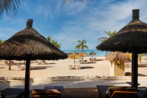 A beach with thatched umbrellas and lounge chairs, a sandy beach, and palm trees. - Royam Hotel (Mbour)