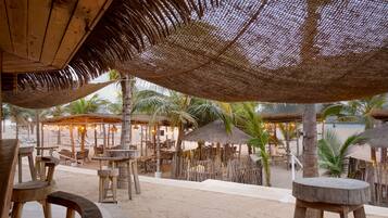 Un espace extérieur aménagé avec des tables et des chaises en bois, des parasols en chaume et des palmiers.