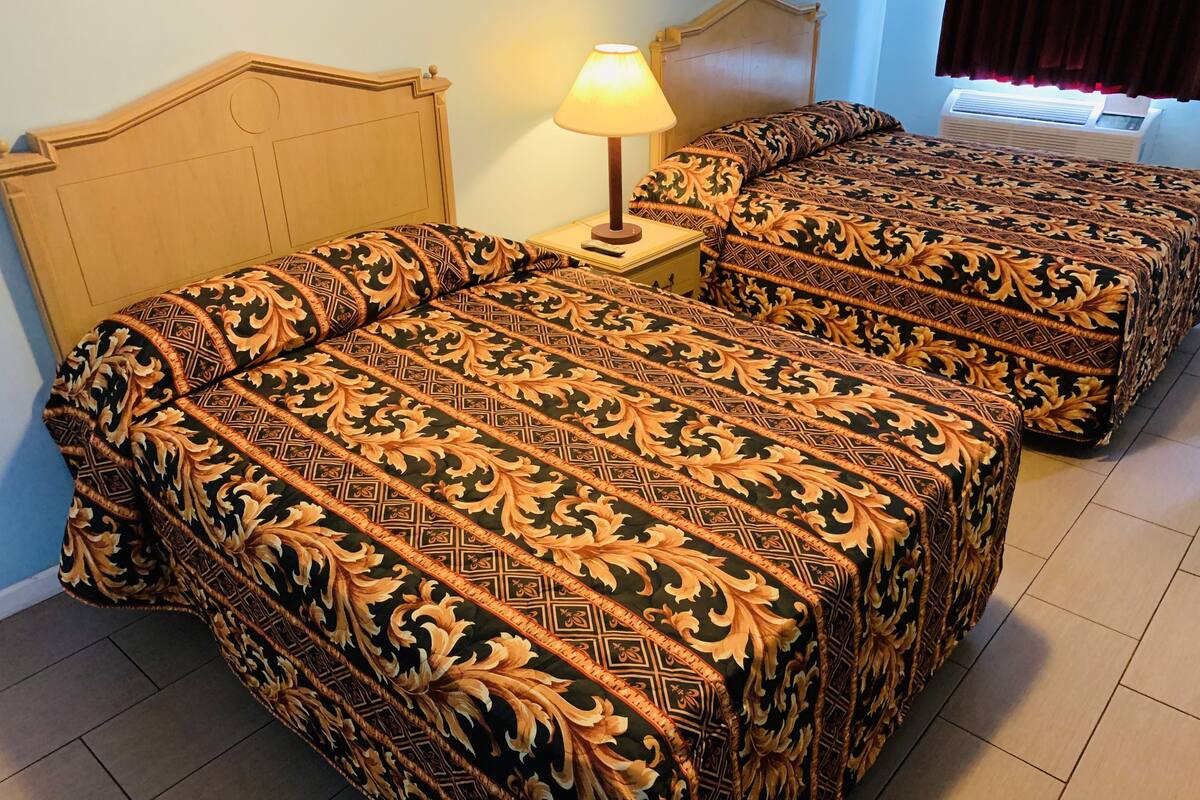 Two beds with matching patterned bedding, a bedside table with a lamp, and a window with curtains.