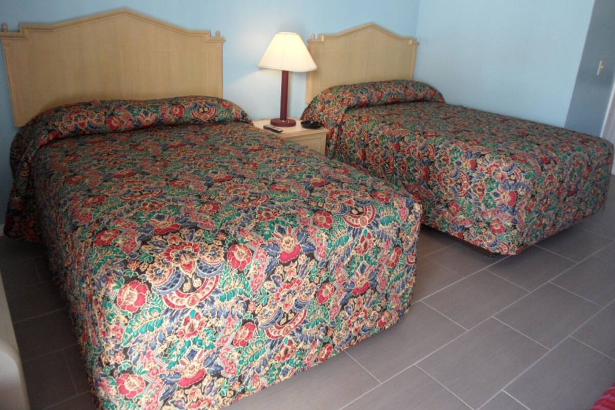 A room with two beds, each with a floral-patterned bedspread, a nightstand with a lamp, and a tiled floor.