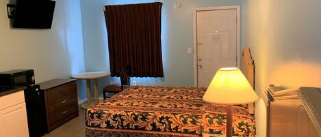 A hotel room with a bed, a desk, a chair, a television, a lamp, and a door.