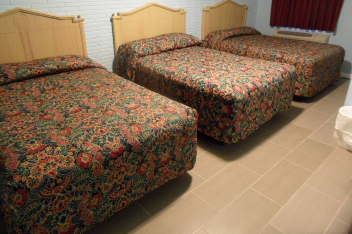 A room with three single beds, each with a wooden headboard and floral-patterned bedding. The room has a white brick wall and a red curtain.