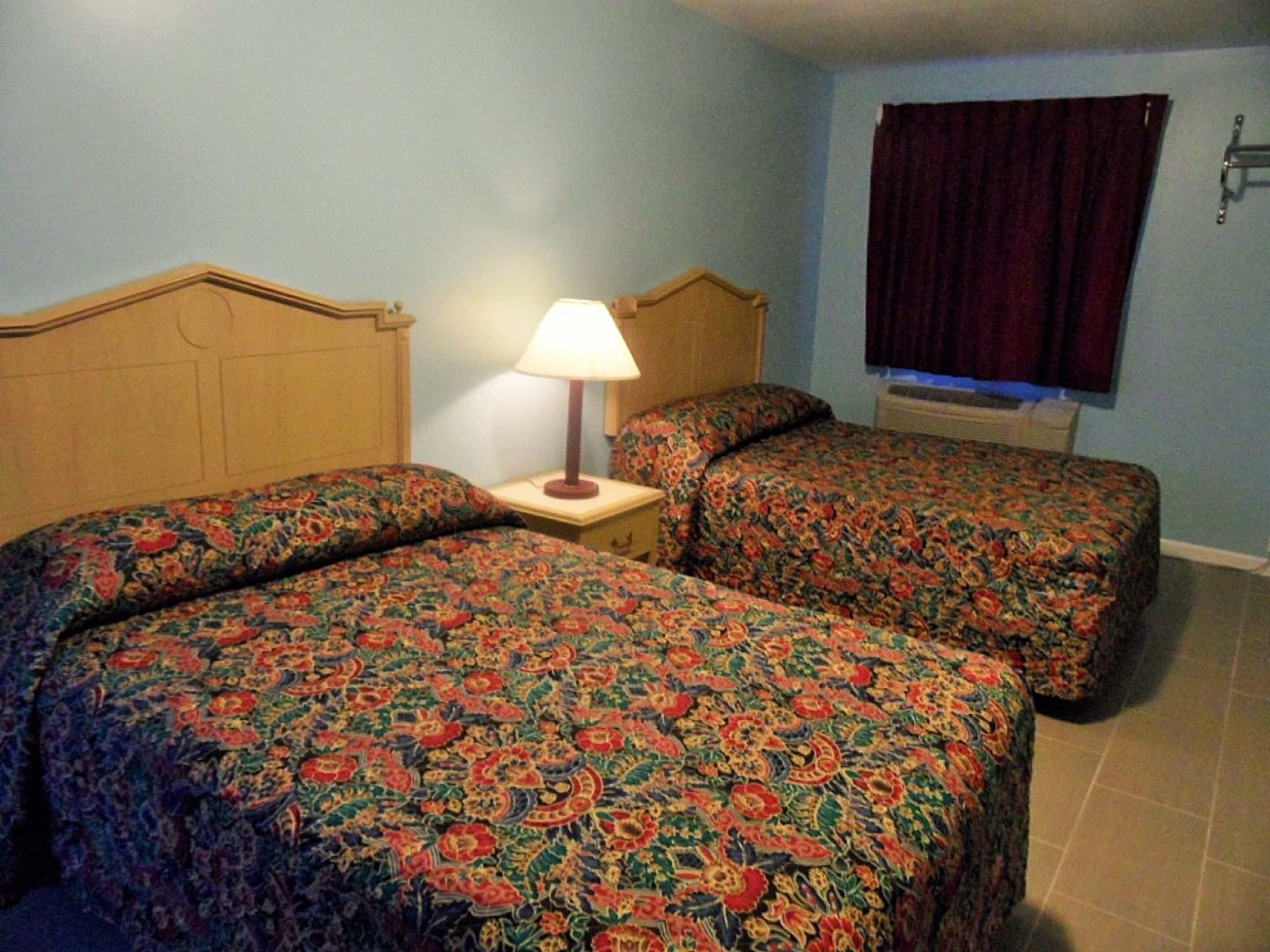 A room with two beds, each with floral-patterned bedding, a nightstand with a lamp, and a window with a curtain.