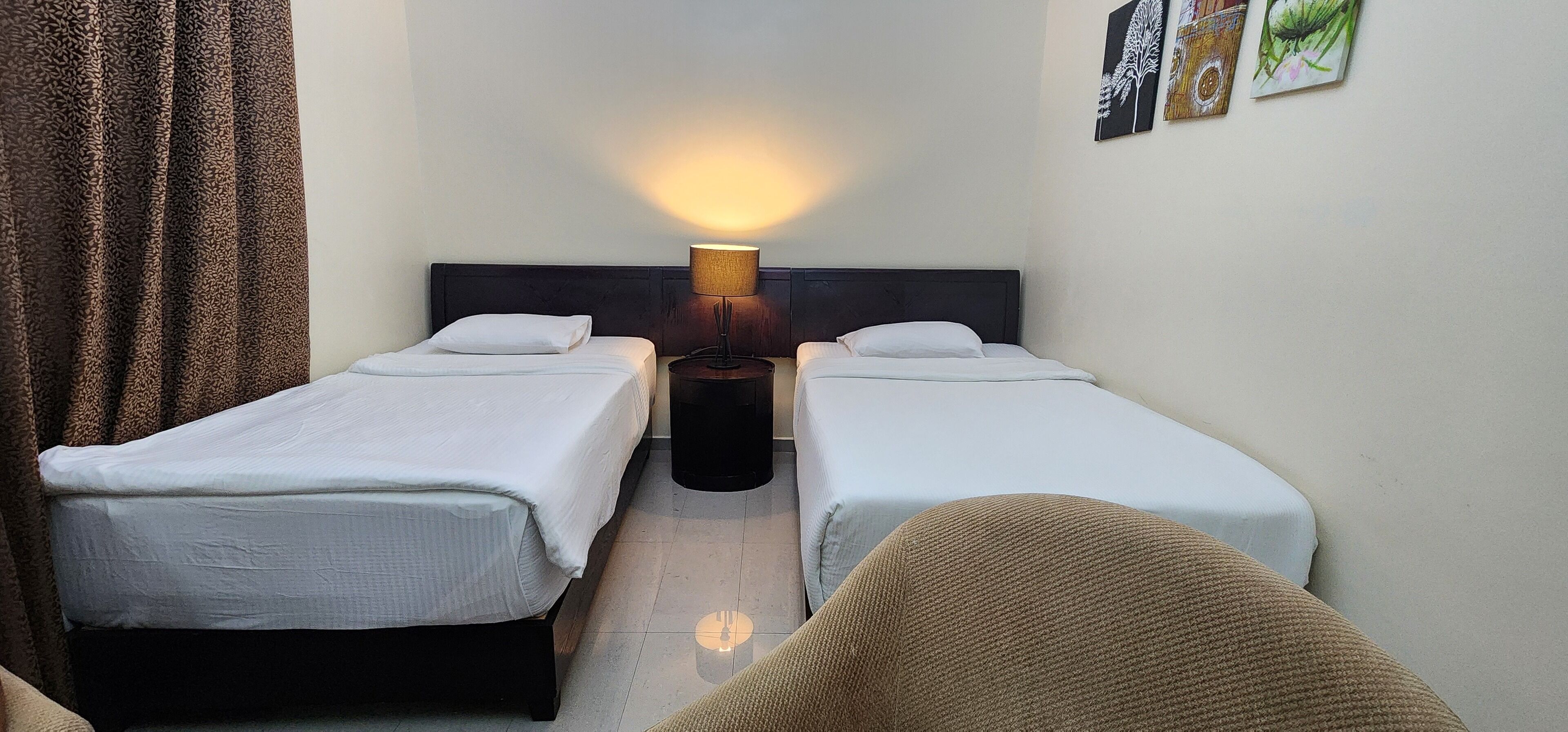 A hotel room with two single beds, a nightstand with a lamp, a chair, and artwork on the wall.