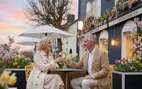 Two people seated at an outdoor table with a drink, surrounded by flowers and a building with 'MOET & CHANDON' signage.