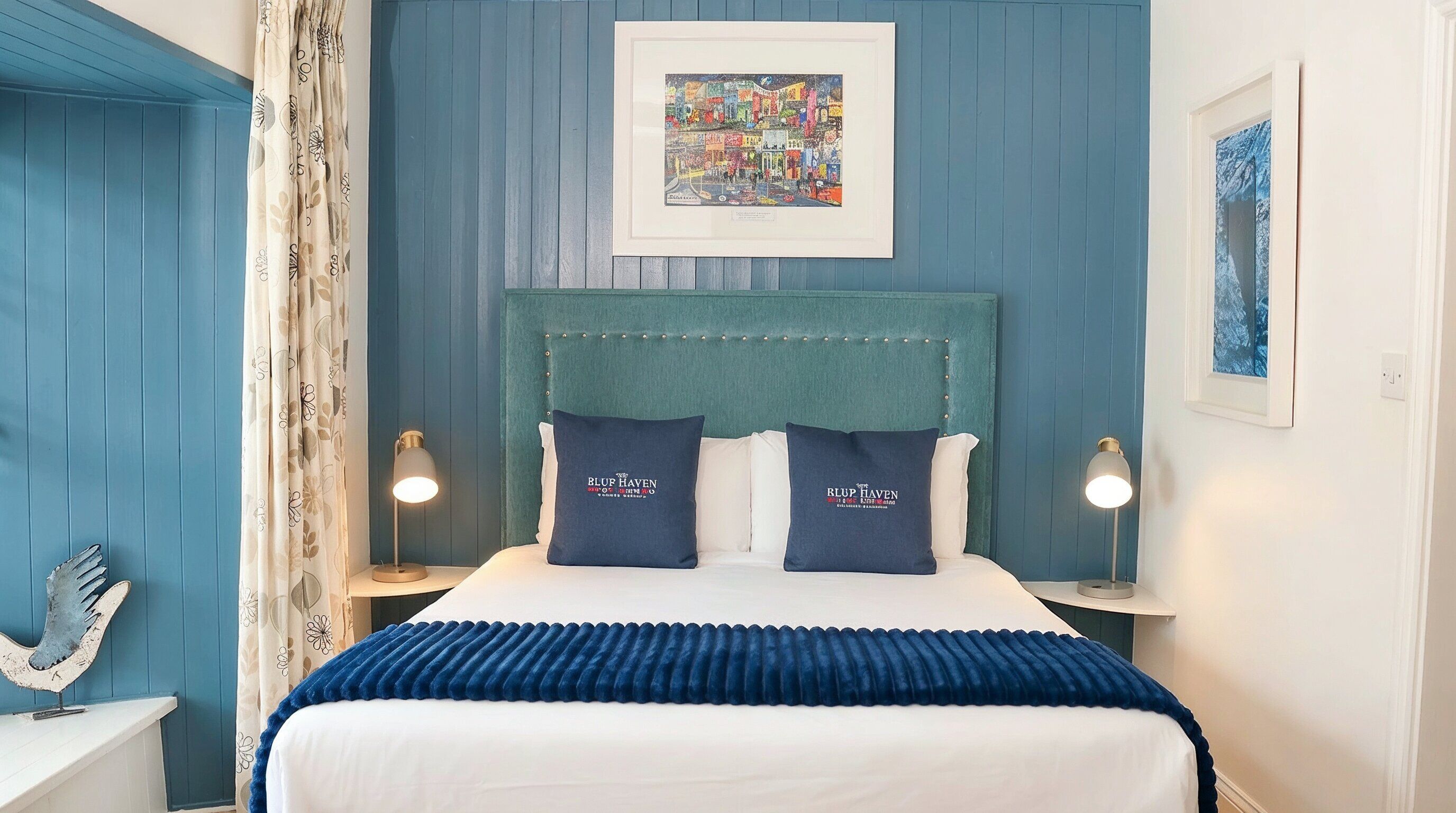 A bedroom with a teal headboard, two pillows with 'BLUF HAVEN' branding, a blue bedspread, and a framed colorful artwork above the bed.
