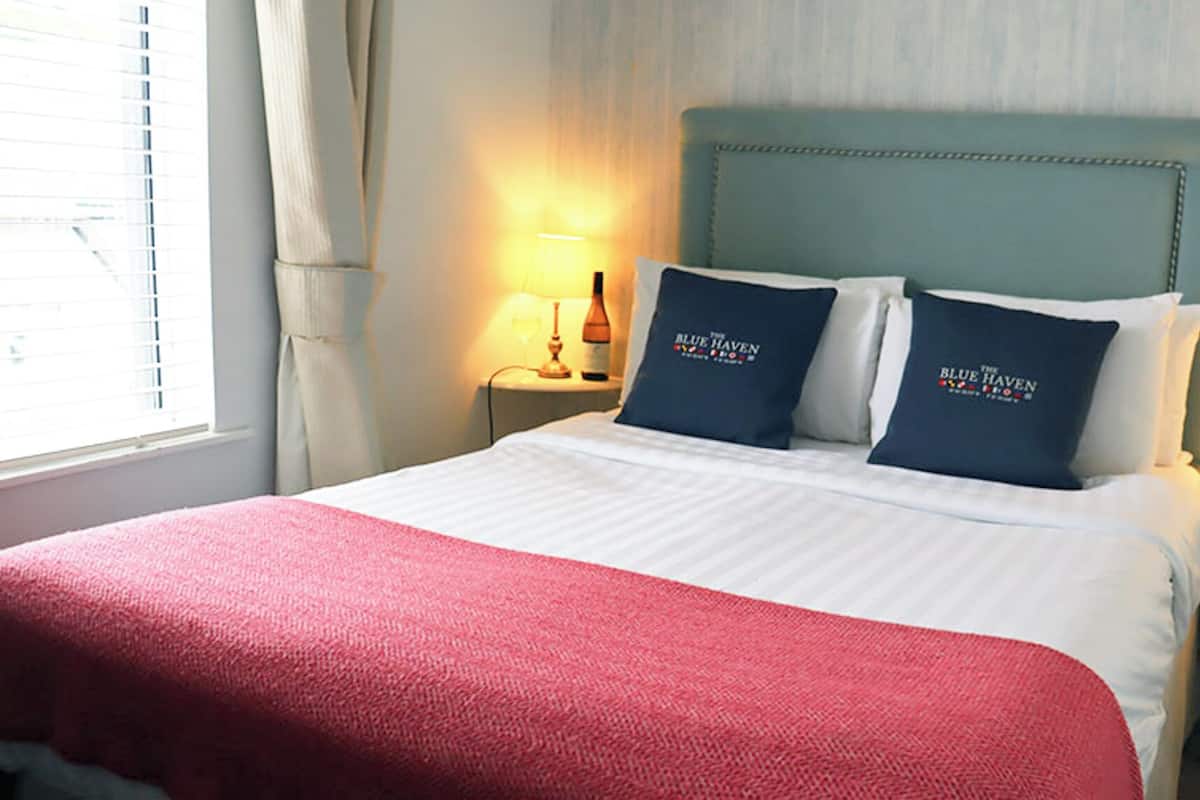 A hotel room with a bed, two pillows with the hotel's logo, a bedside table with a lamp, and a window with curtains.