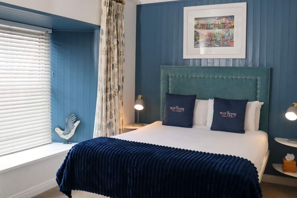 A bedroom with a bed, two bedside lamps, a framed picture on the wall, and a window with blinds.