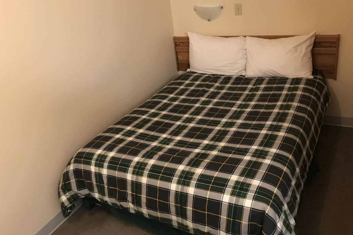 Un lit bien fait, recouvert d’une couverture à carreaux et agrémenté de deux oreillers blancs, avec une tête de lit en bois et un luminaire fixé au mur.