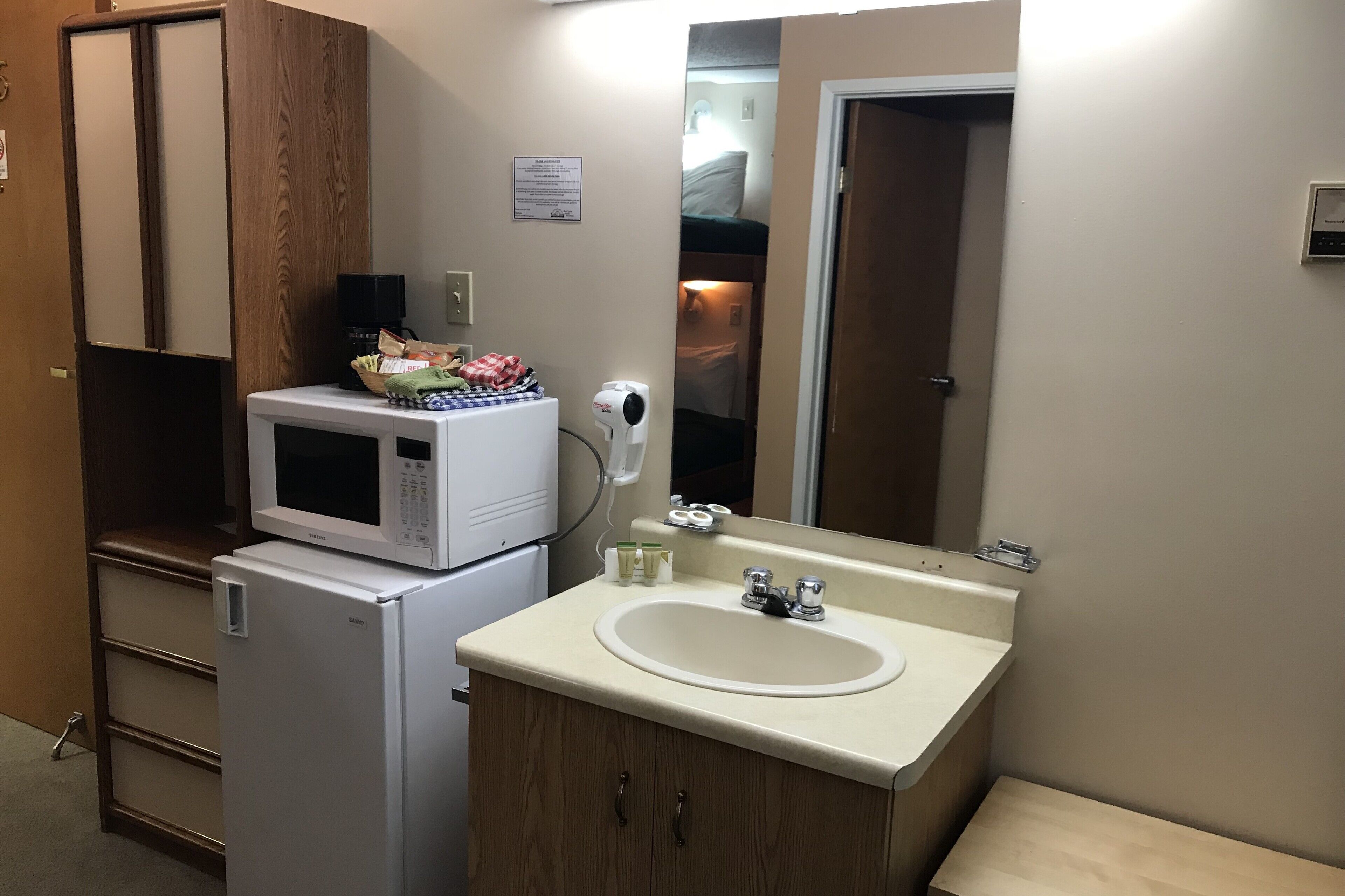 A compact hotel bathroom with a white sink, a microwave on a small refrigerator, a hand dryer, and a wooden vanity.