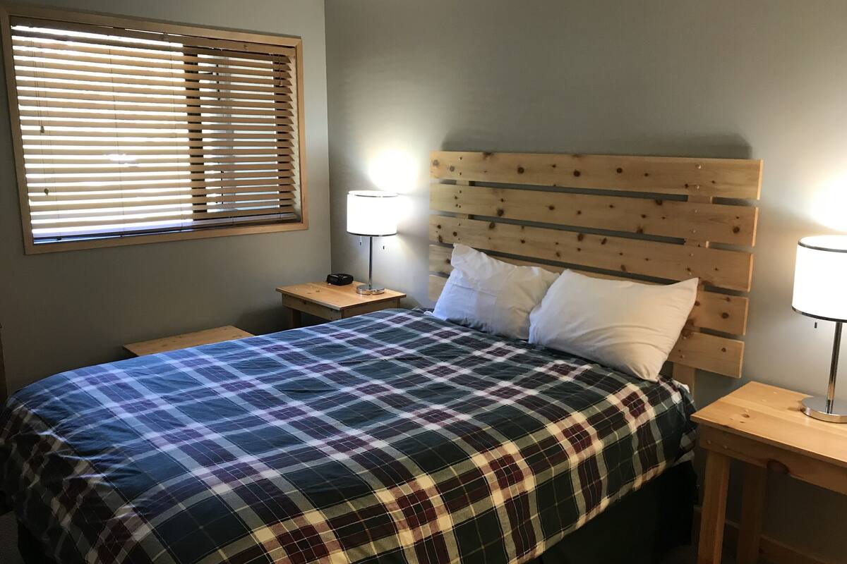 Une chambre à coucher avec une couvre-lit à carreaux, une tête de lit en bois, deux lampes de chevet et une fenêtre avec des stores.