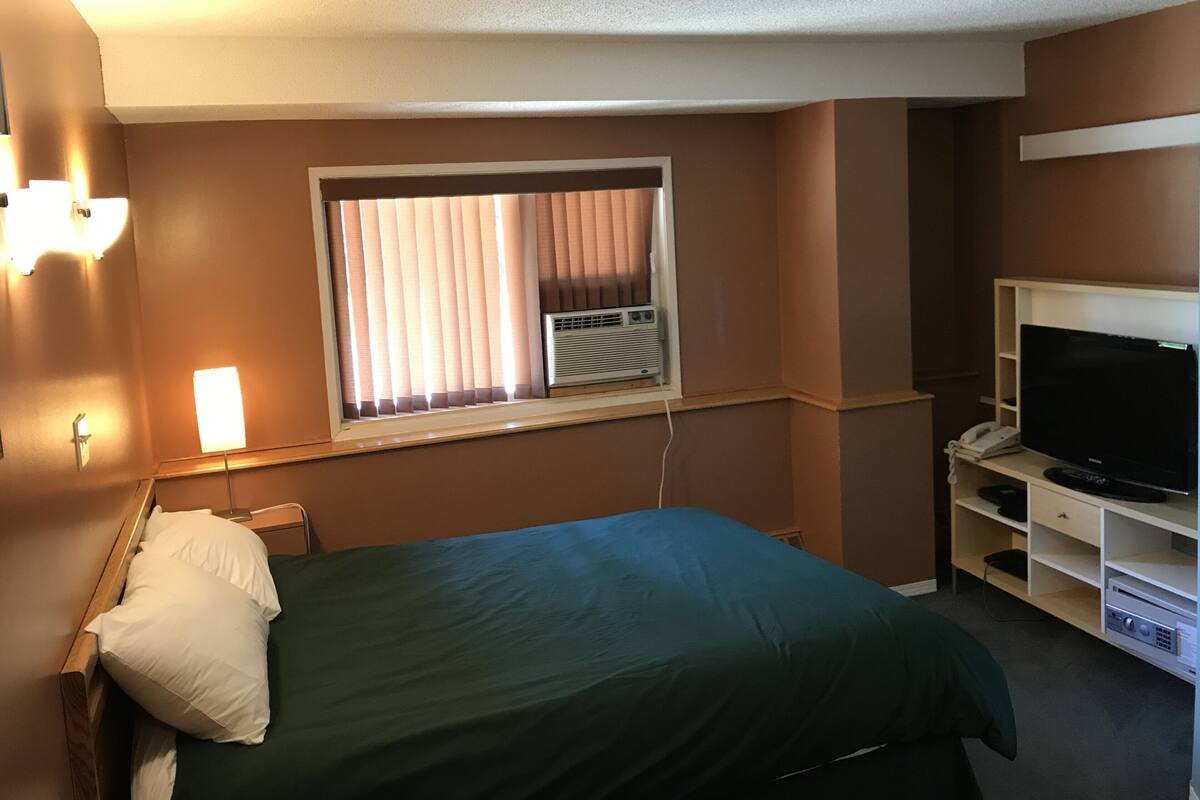 Une chambre d’hôtel équipée d’un lit, d’une télévision, d’une fenêtre avec stores et d’un climatiseur fixé au mur.