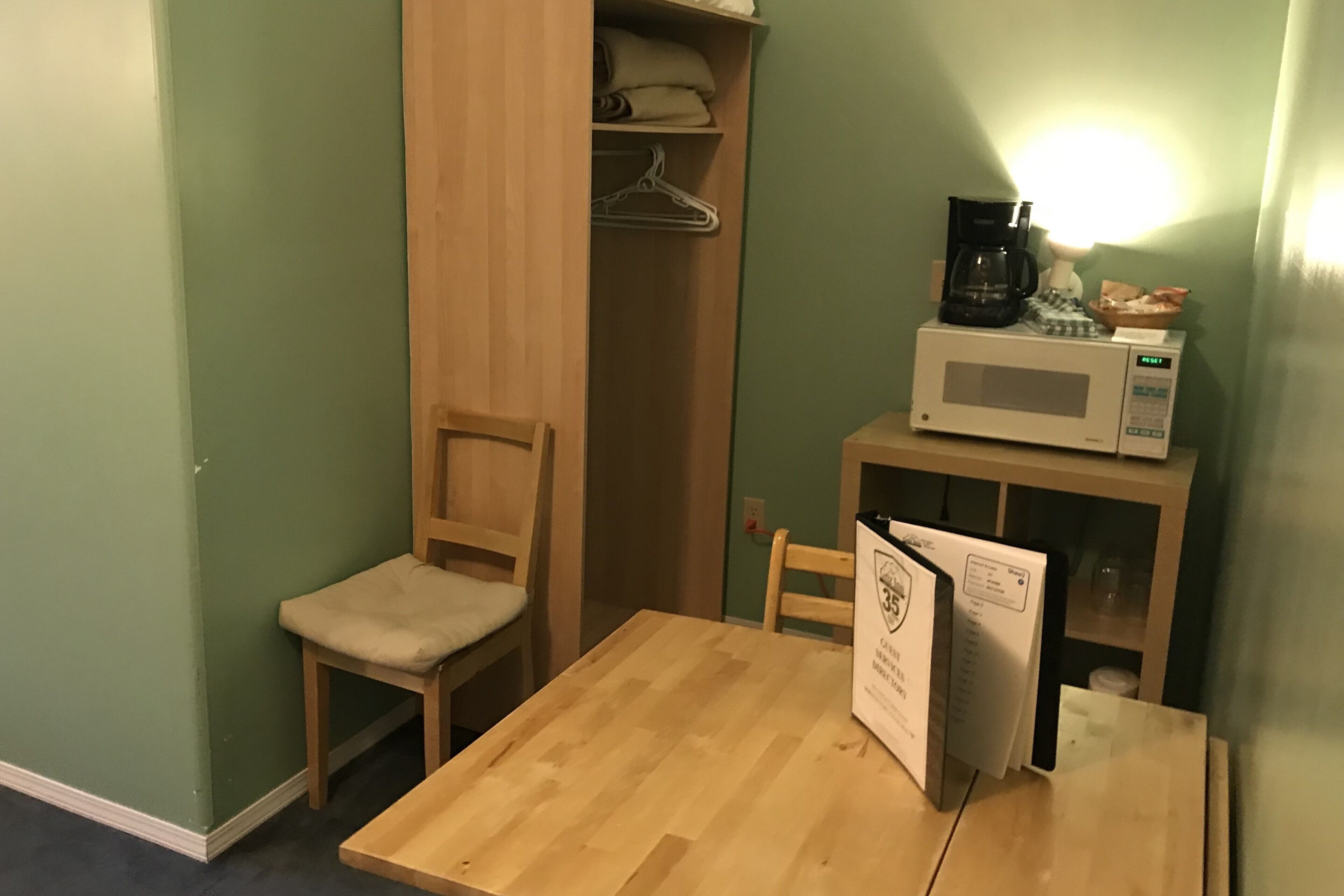 A small room with a wooden table and chairs, a microwave, a coffee maker, and a closet with hanging clothes.