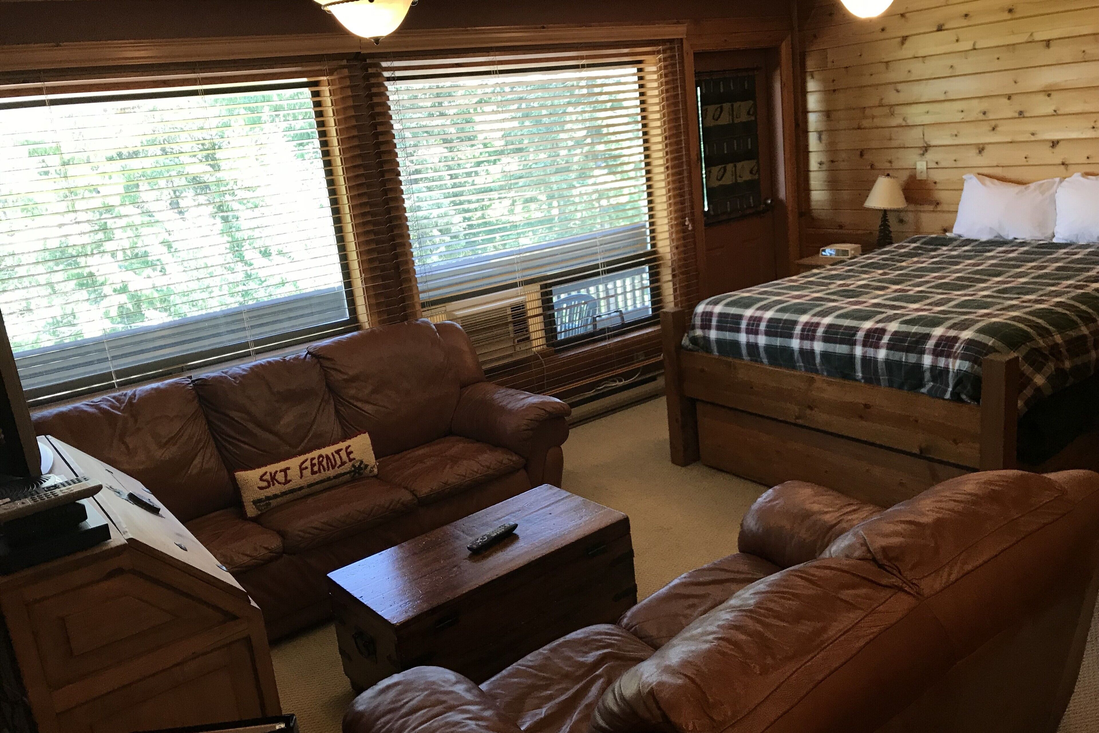 Una habitación acogedora con un sofá de cuero marrón, una cama de madera con colcha escocesa, un pequeño escritorio de madera con lámpara y una ventana con persianas.