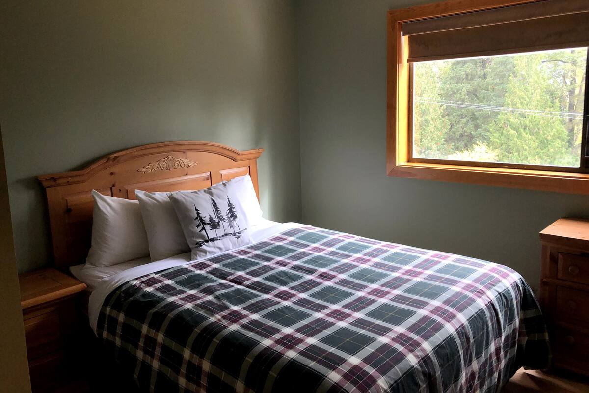 Une chambre à coucher avec une tête de lit en bois, une couvre-lit à carreaux et une fenêtre donnant sur les arbres.