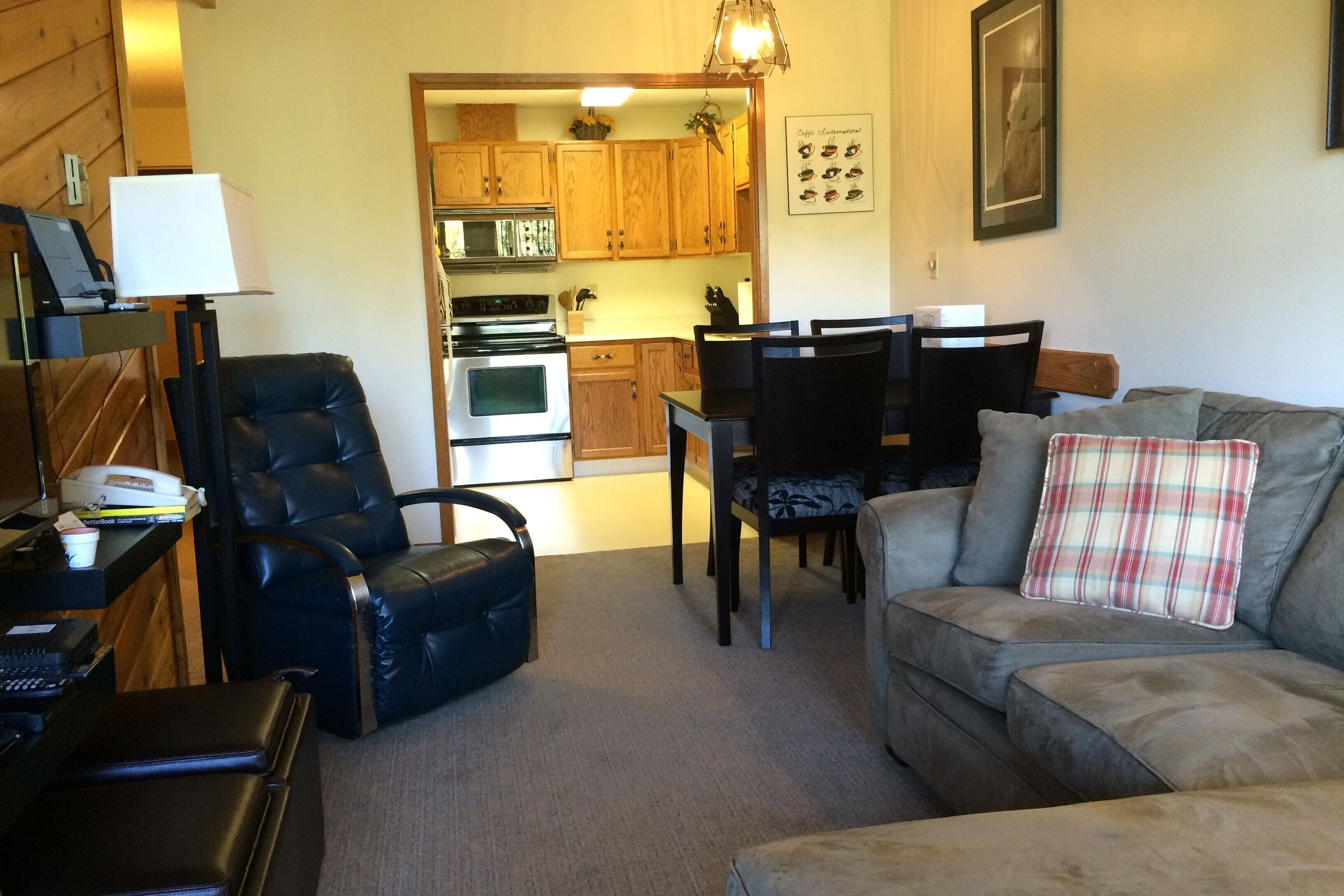 A living room with a sofa, a black leather chair, a dining table with chairs, and a kitchen area with wooden cabinets and appliances.