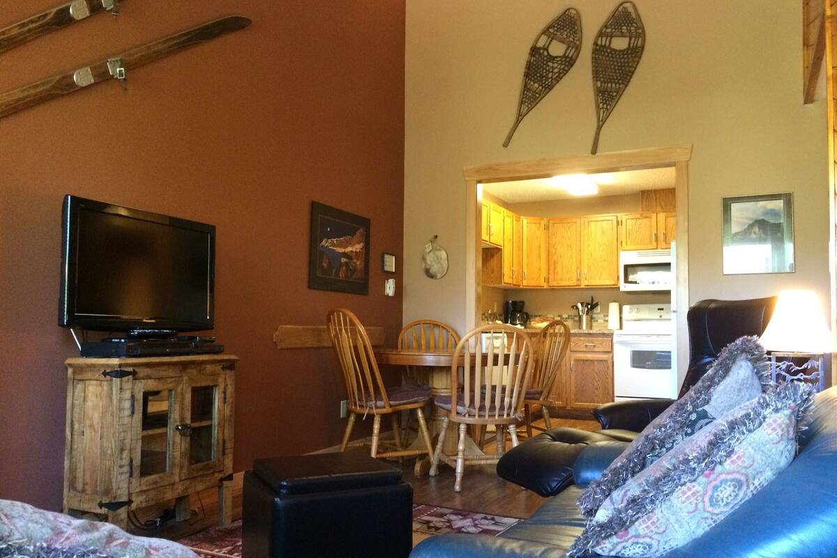 Un salon avec un téléviseur posé sur un meuble en bois, un coin repas avec des chaises en bois, et une cuisine visible à travers une porte ouverte.