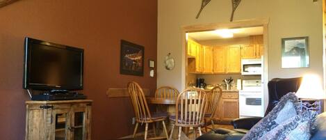A living room with a television on a wooden cabinet, a dining area with wooden chairs, and a kitchen visible through an open doorway.