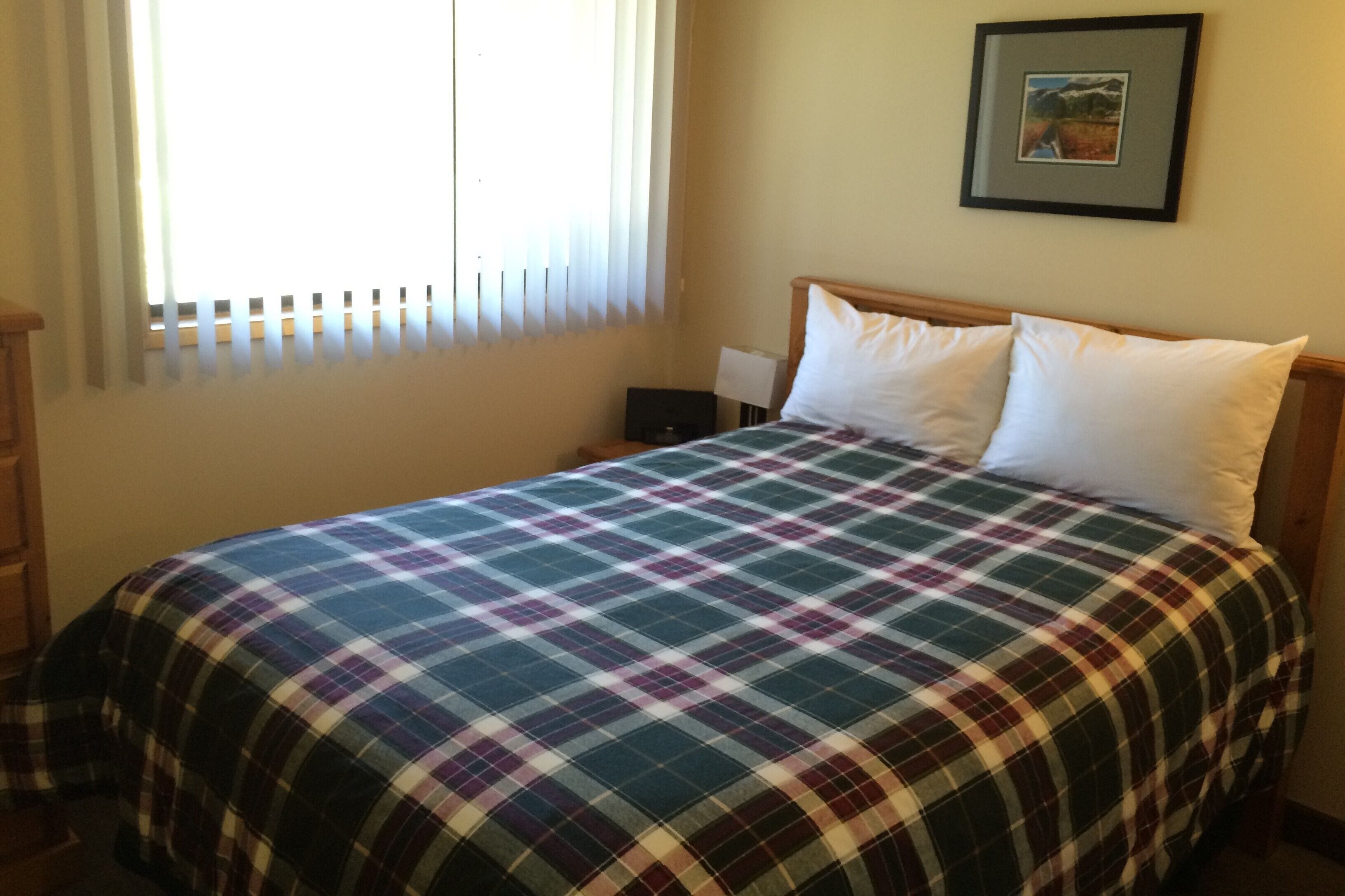 A neatly made bed with a plaid bedspread, a bedside table with a lamp, and a framed picture on the wall.