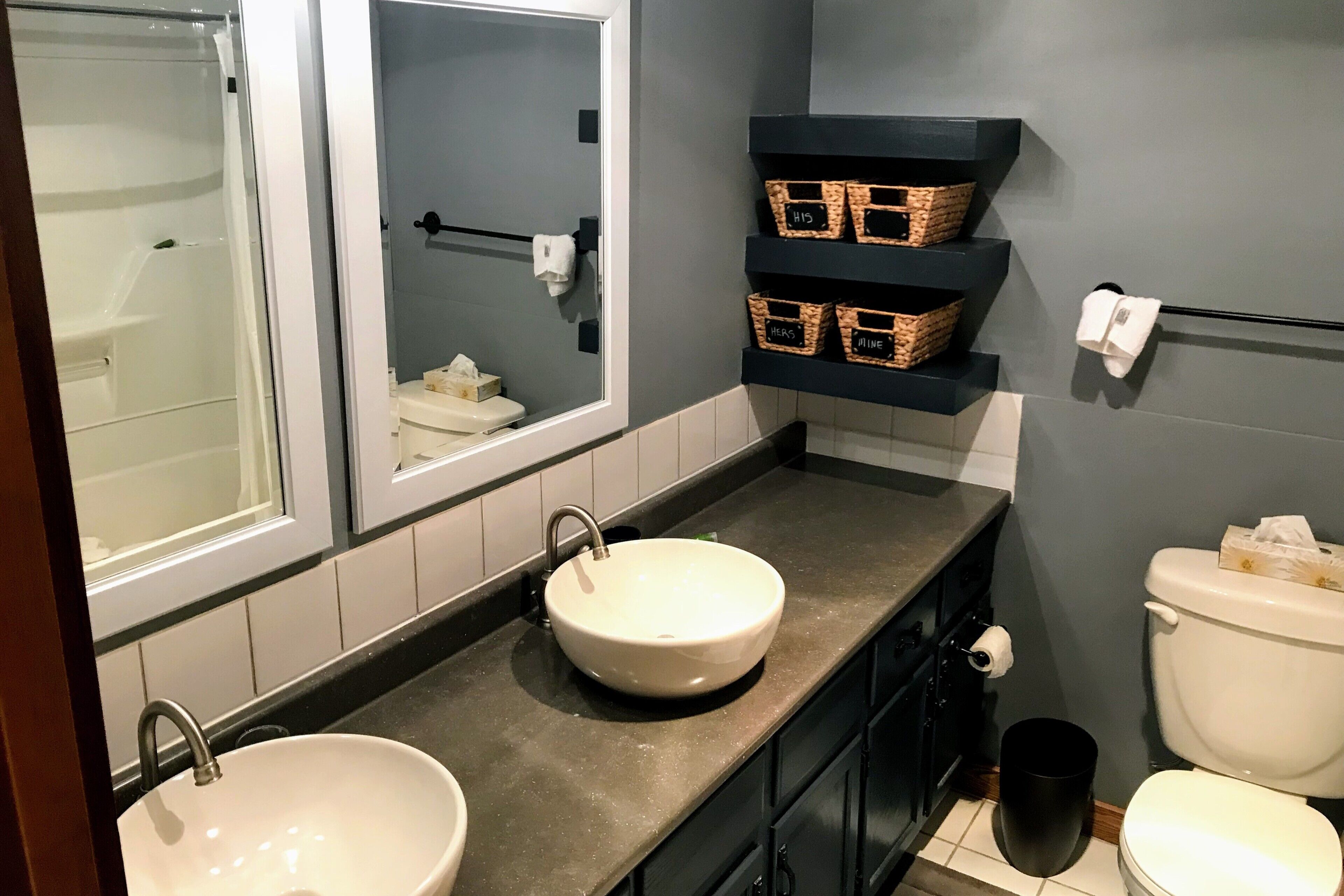 Ein Badezimmer mit zwei Waschbecken, einer Toilette und einer Dusche. Auf Regalen stehen Korbwannen und es gibt einen Handtuchhalter.