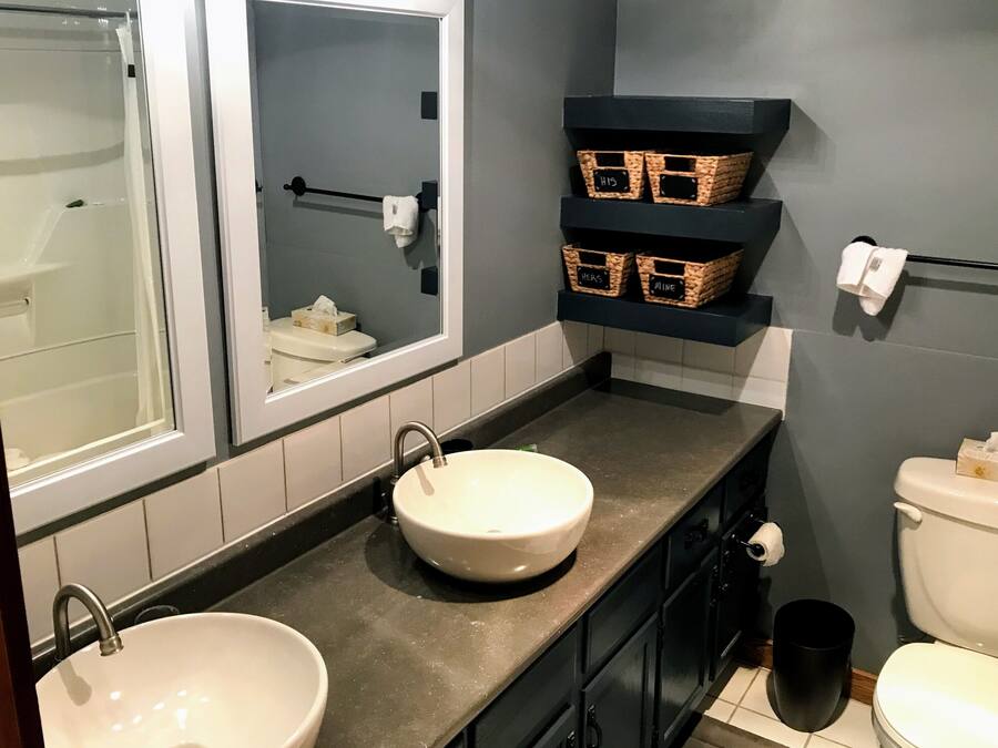 Ein Badezimmer mit zwei Waschbecken, einer Toilette und einer Dusche. Auf Regalen stehen Korbwannen und es gibt einen Handtuchhalter.