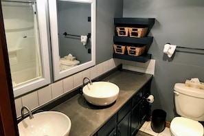 A bathroom with two sinks, a toilet, and a shower. There are wicker baskets on shelves and a towel rack.