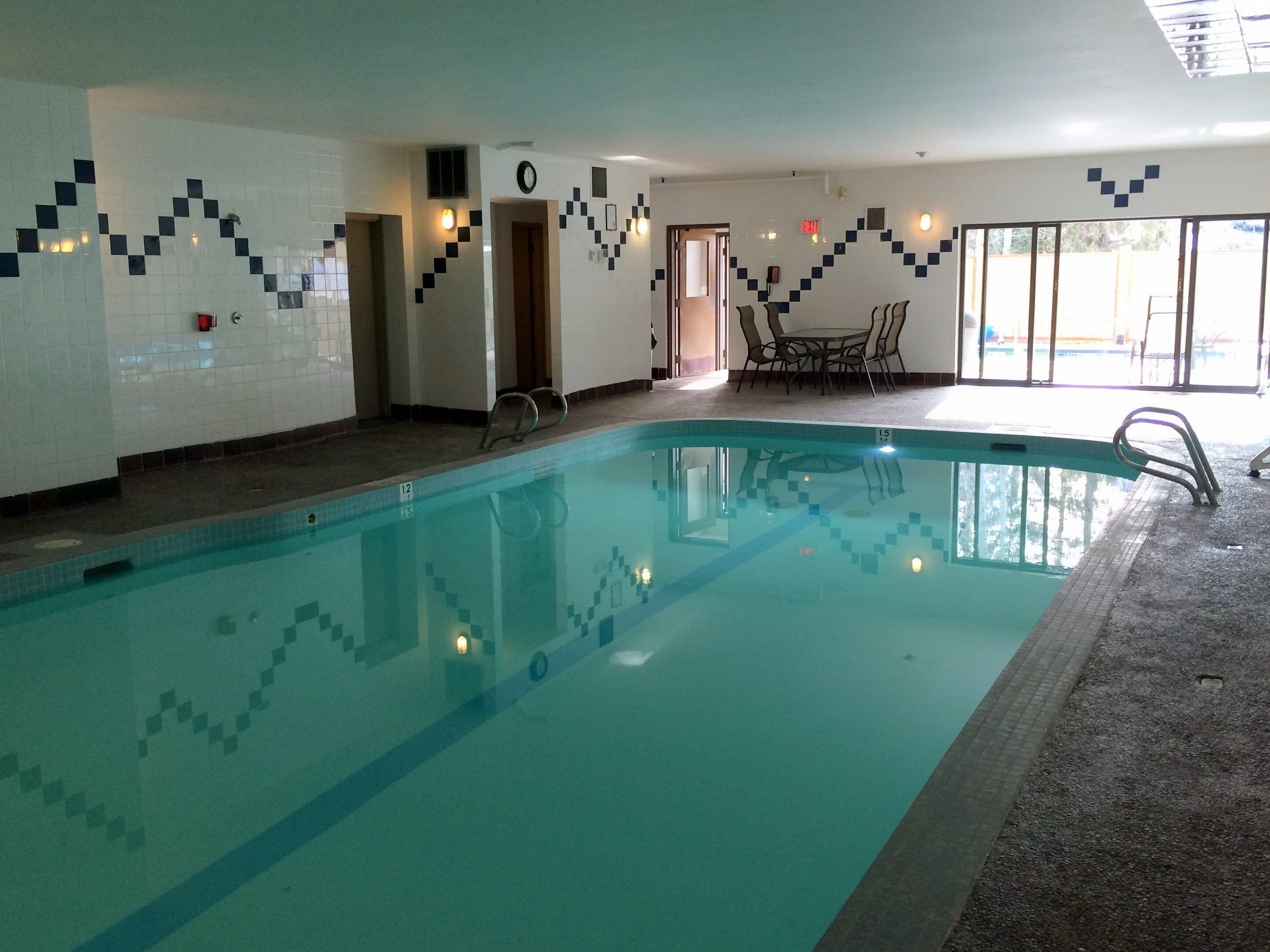 Una piscina interior con agua cristalina, paredes de azulejos y una zona de estar con sillas y mesas.