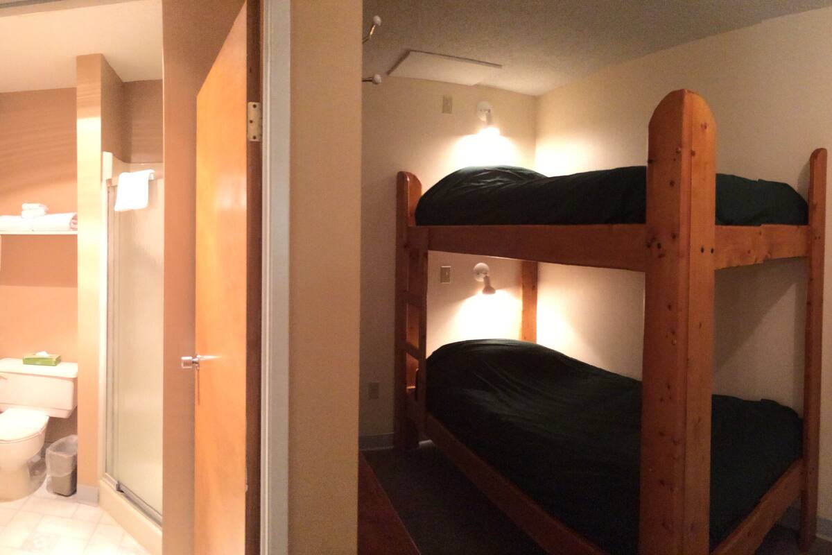 Une petite pièce compacte avec un lit superposé et un lit simple. On aperçoit une salle de bain sur la gauche, avec des toilettes et une douche.