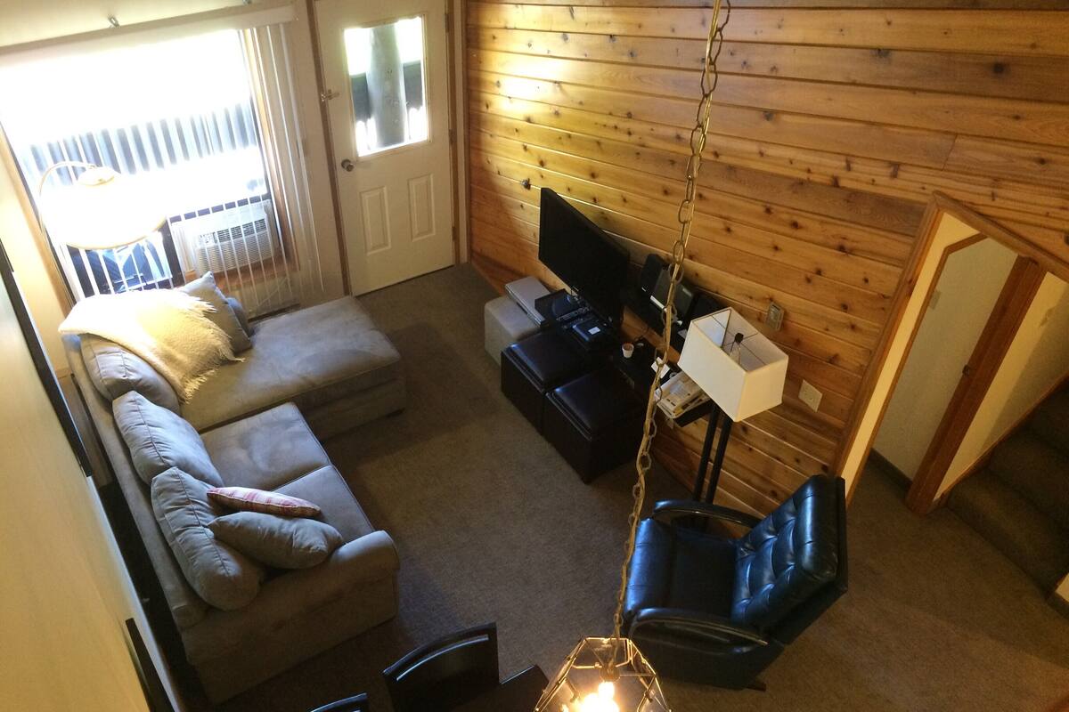 Un salon chaleureux avec un canapé, un fauteuil en cuir noir, un téléviseur et un luminaire suspendu.