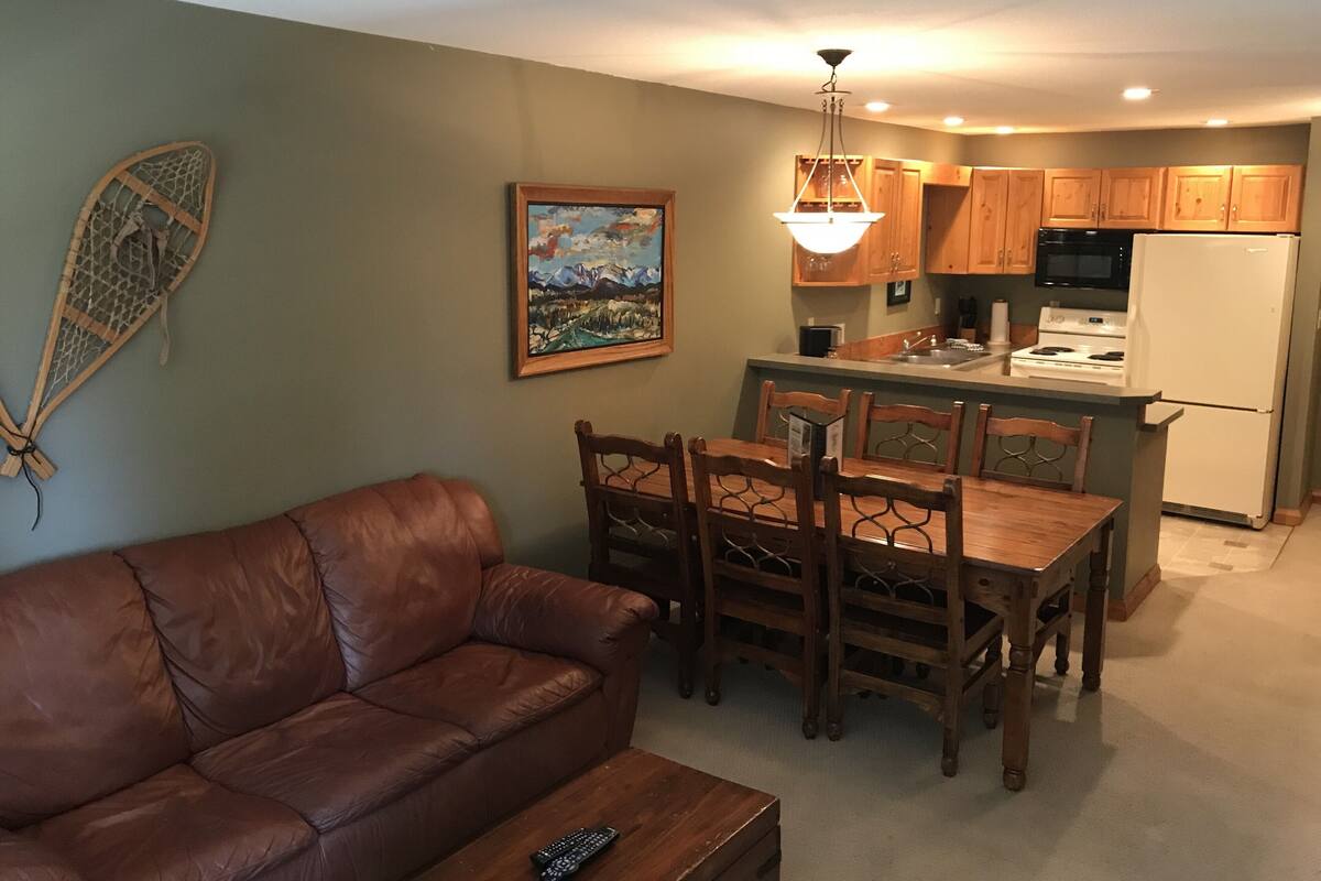 Un salon avec un canapé en cuir marron, une table à manger en bois avec des chaises, un coin cuisine avec des meubles en bois, un réfrigérateur blanc et un tableau accroché au mur.