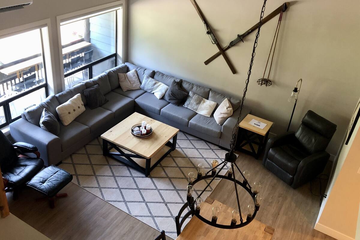 Un salon moderne avec un canapé d’angle, une table basse en bois, un fauteuil en cuir noir et un luminaire suspendu.